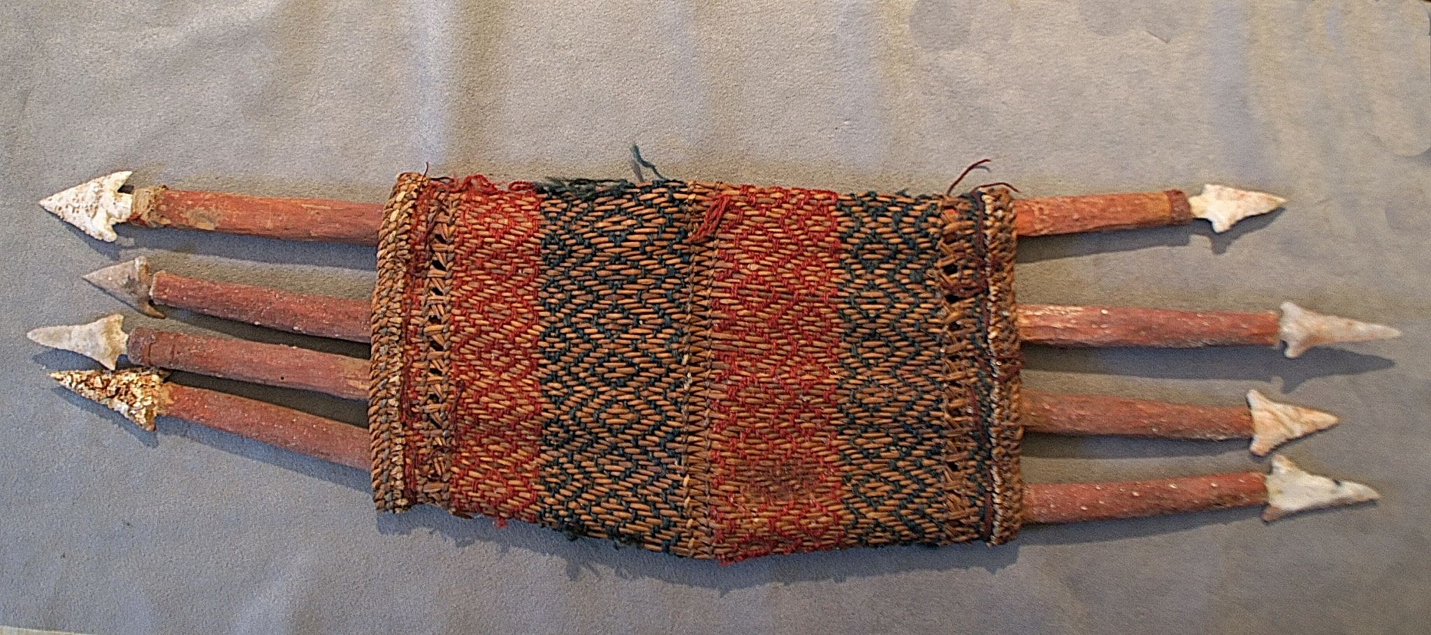 Chile, Arica Harpoon Quiver With Eight Harpoons
This decorative fiber quiver is woven with dyed red and green reeds, and is intact with its original wood harpoon points and napped stone arrows.  The wood harpoon points would have been set into a longer throwing stick; the point releases when the harpoon is retrieved. The quiver, also known as a porta harpoon (harpoon carrier) holds eight points.  It was used by the fisherman to hold ammunition on extended fishing trips.  Junius Bird describes and illustrates these porta harpoons in "EXCAVATIONS IN NORTHERN CHILE," 1943, pg. 223.  The harpoons are painted red, which was ceremonial and indicates that they were probably for the afterlife
Media: Wood
Dimensions: Length: 23" Width: 7"
n6041