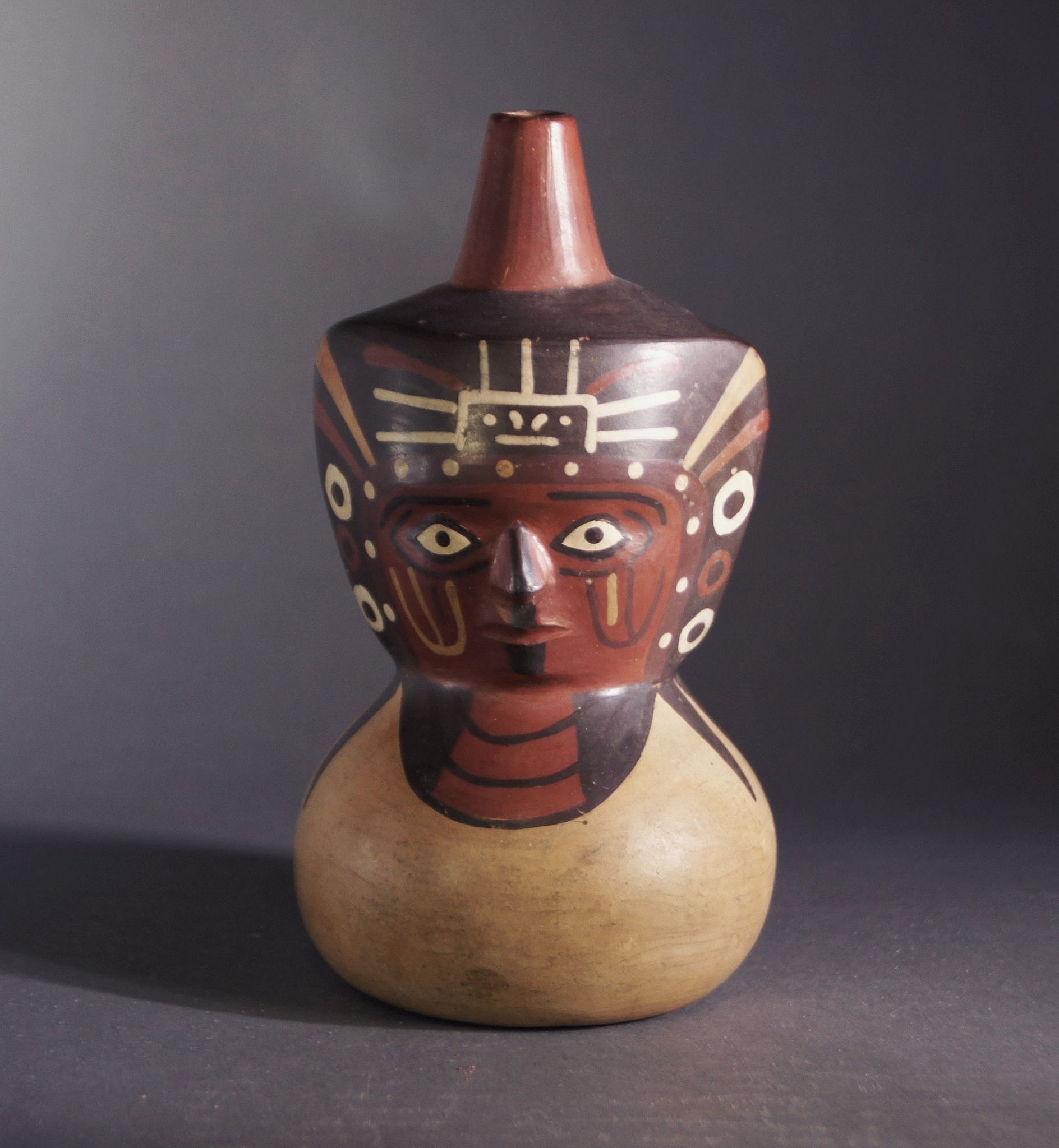 Peru, Wari Single Spout Figural Bottle with Elaborate Facial Decoration
This effigy vessel is painted with an individual wearing a diadem on his headdress and as well as a triple-strand necklace, indicating that he is a shaman or other important personage. He wears a black head covering, which is depicted draped around the shoulders.  Ex. collection Anton Roeckl, Munich. T/L tested in Berlin.
Media: Ceramic
Dimensions: 6 in.
Price Upon Request
94114A