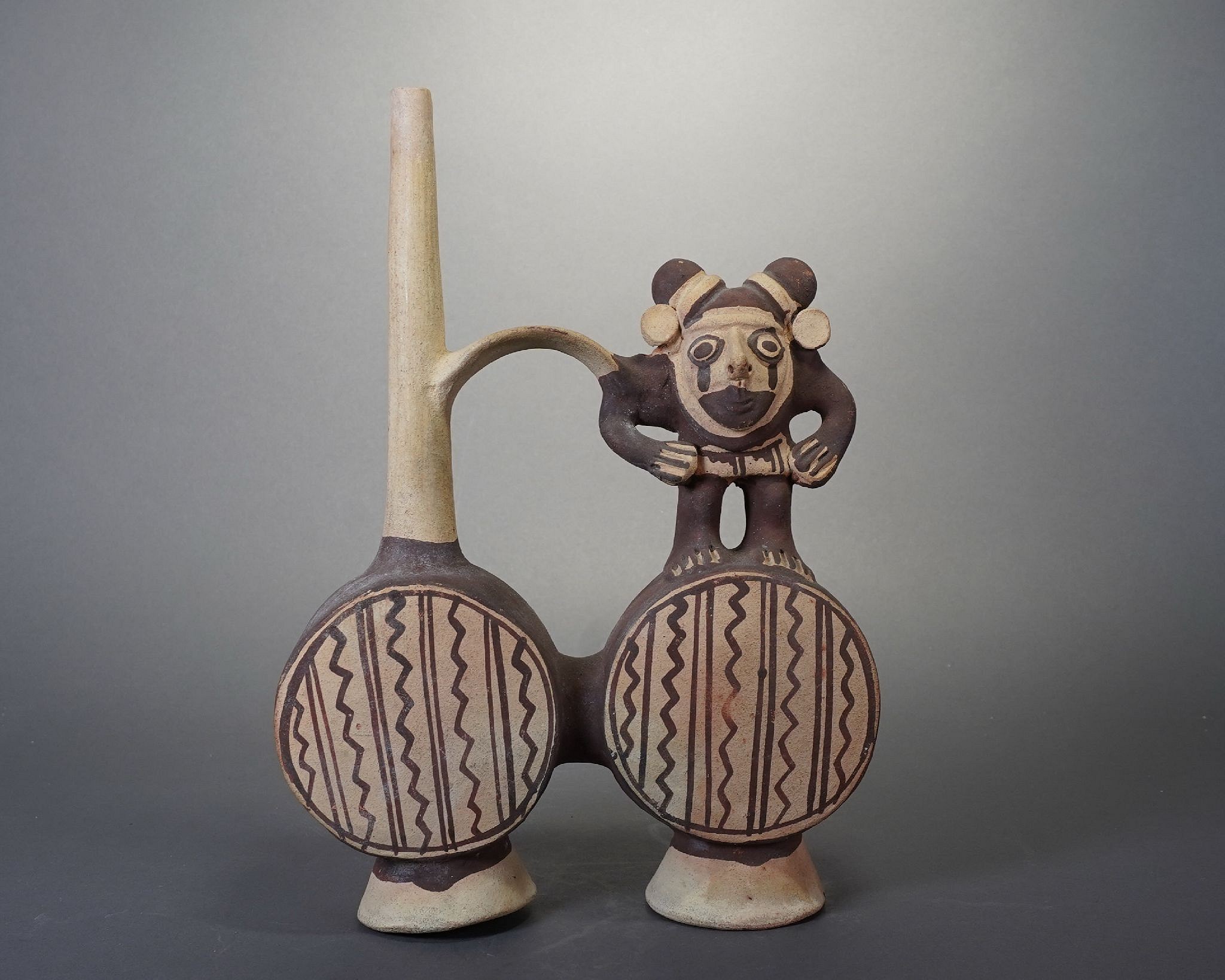 Peru, Chancay Double-Chambered Whistling Vessel with Animal Impersonator
The Chancay people were known for their ceramic workshops, where series of similar items were produced in groups.  This bridge-spout vessel has two chambers and is painted with alternating straight and wavy lines.  The figure sitting atop the vessel is an Animal Impersonator of a monkey.  “Animal Impersonator” is a term used by Pre-Colombian scholars to refer to a shaman who dresses as a certain animal to embody the mythological characteristics associated with that animal.  Monkeys, who dwell at the top of the forest canopy in the Amazon, are associated with ancestral secrets in Andean mythology.  This shaman has a hairstyle arranged in two coils to resemble monkey ears, as well as white ear spools.  He is hunched over and holds a weaving in his arms. 
This vessel may have been used to pour a psychoactive brew as part of a shamanic rite.   When brew was poured, the air moved from one chamber to the other, creating a whistling sound.  The tone of the whistle was created by the specific shape of the resonance cavity.  Whistling sounds would have enhanced the shamanic ritual.  According to author Daniel K. Stat’s article Ancient Sounds: The Whistling Vessels of Peru, “the psycho-acoustical effects appear to be created by low frequencies or difference tones, or possibly the interaction of harmonic principles, which are produced when several vessels from a specific culture are played simultaneously.”  
This vessel would also have been intended for its owner for use in the afterlife. 
Media: Ceramic
Dimensions: Height: 10 1/2" x Width: 7"
$1,400
n9047