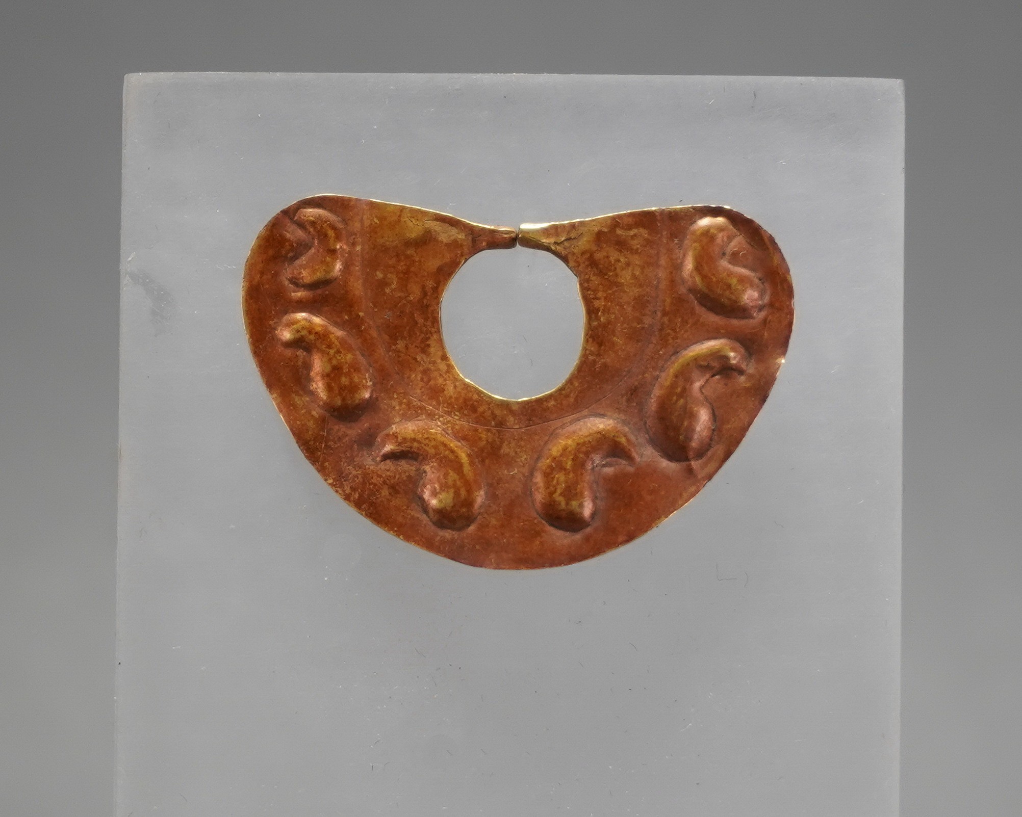 Peru, Moche Gold Nose Ornament with 6 Embossed Uculla Fruit
The Uculla fruit was an anti-coagulant to thin prisoners' blood for ritual ceremonies.  The patina contains ferric oxide from the soil in the vicinity of the burial site, which gives the gold ornament its beautiful red patina.  This piece was hammered from a soft sheet of pure gold.  Small gold ornaments such as this required excellent craftsmanship to execute.
n9056